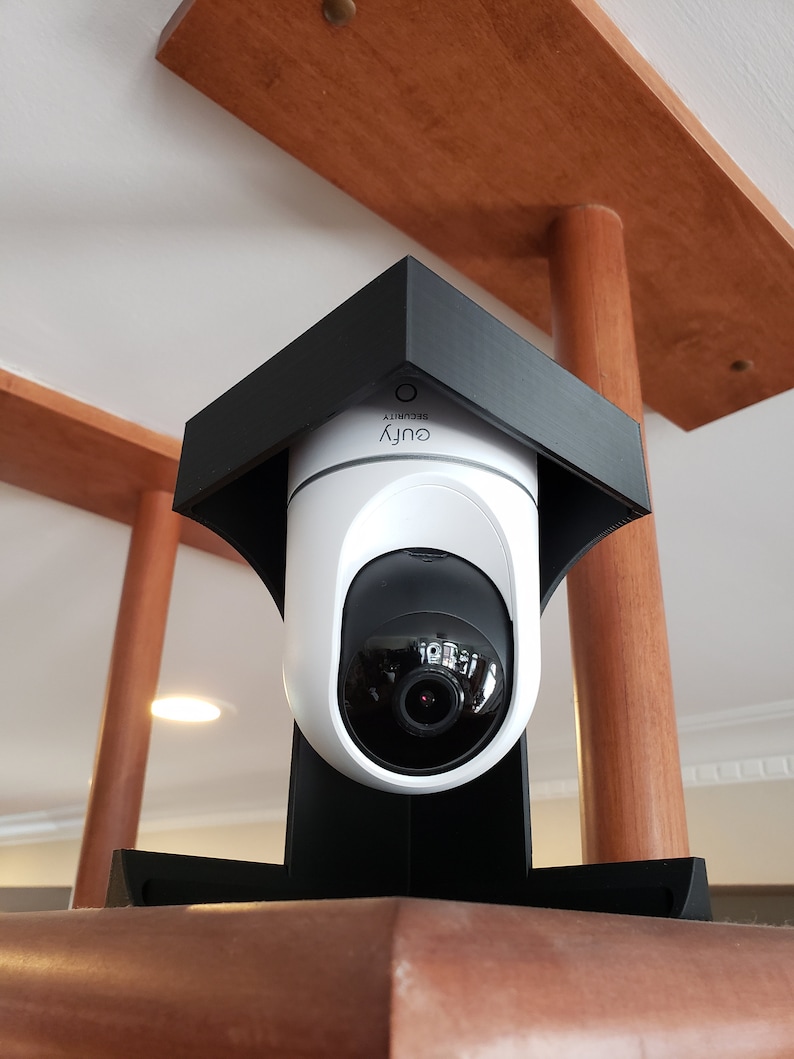 May include: A white and black security camera with the word "eufy SECURITY" printed on it. The camera is mounted in a black housing and is positioned under a wooden structure. The camera lens is visible.