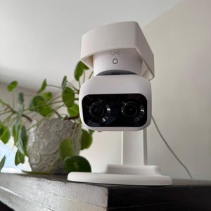 Op de afbeelding: Een witte beveiligingscamera met een witte zonnekap erboven. De camera heeft twee zwarte lenzen en de tekst "Outly" is op de zonnekap gedrukt. De camera is gemonteerd op een witte standaard en staat op een donkerhouten plank.