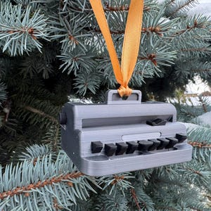 May include: A gray typewriter ornament with black keys and a handle, hanging from a gold ribbon on a blue-green evergreen tree. The ornament has a vintage aesthetic, perfect for holiday decor.