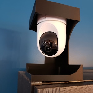 May include: A white and black security camera with the word "eufy" printed on it. The camera is mounted on a black bracket. The camera is positioned on top of a dark brown cabinet against a blue wall.