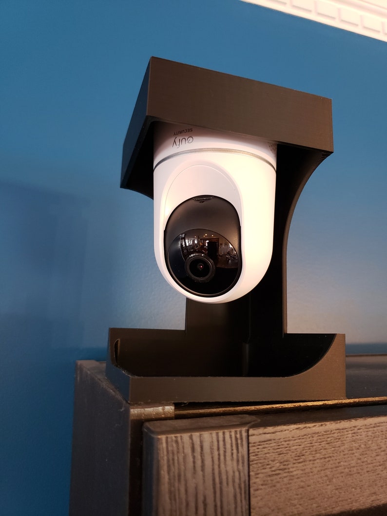 May include: A white and black security camera with the word "eufy" printed on it. The camera is mounted on a black bracket. The camera is positioned on top of a dark brown cabinet against a blue wall.