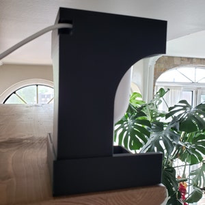 May include: A black, contemporary lamp with a rectangular base and a curved top. The lamp is placed on a wooden surface, with a white cable extending from the top. A large green plant is visible in the background.