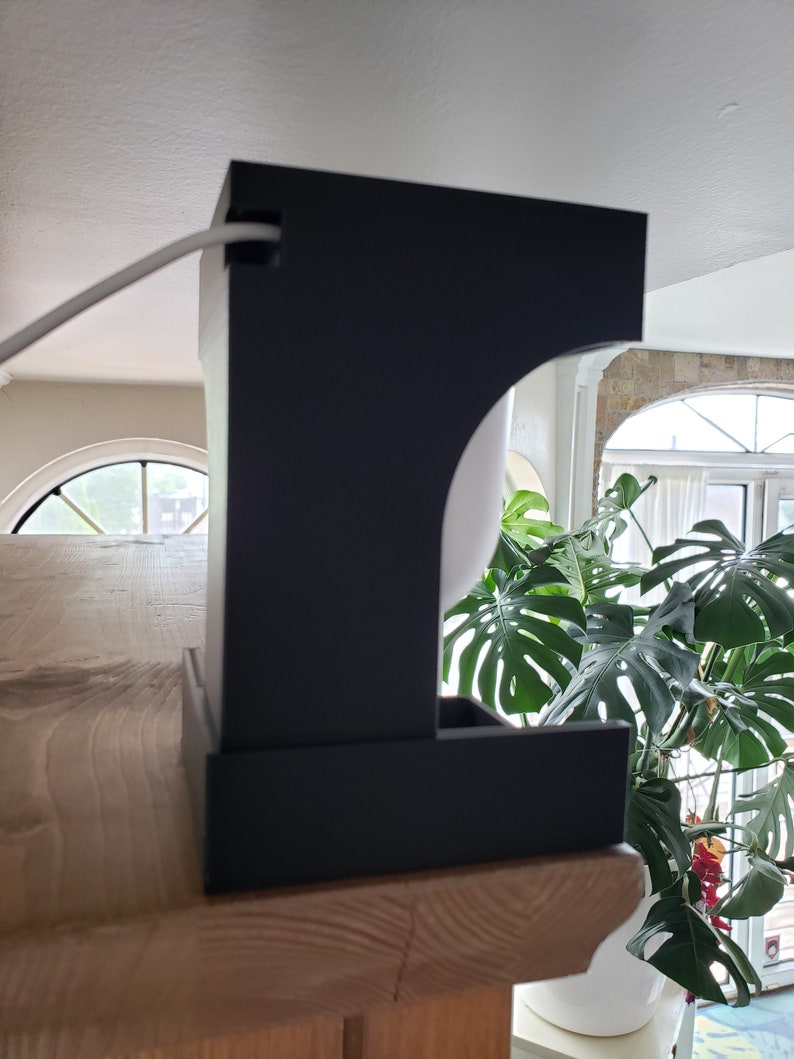 May include: A black, contemporary lamp with a rectangular base and a curved top. The lamp is placed on a wooden surface, with a white cable extending from the top. A large green plant is visible in the background.