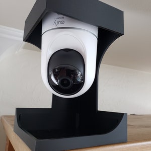 May include: A white and black security camera with the brand name "eufy SECURITY" printed on it. The camera is mounted in a black bracket. The bracket is on a wooden surface.