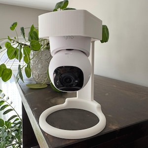 May include: White security camera mounted on a white stand with a white cover. The camera is facing forward and the stand is on a dark brown surface.