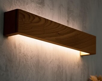 May include: A rectangular wooden wall sconce with a warm, inviting glow. The sconce is made of light brown wood and has a recessed light source that emits a soft, diffused light. The fixture is mounted on a textured gray wall.