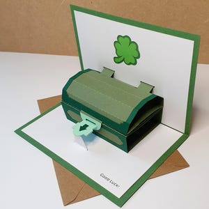 May include: A pop-up greeting card with a green treasure chest design, a shamrock, and the words "Good Luck!". The card is open, displaying the 3D chest. The card is on a white surface.