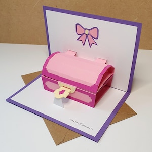 May include: A pop-up birthday card featuring a pink treasure chest design with a bow illustration. The card has a purple border and the words "Happy Birthday!" are printed at the bottom. The card is displayed with a brown envelope.