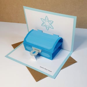 May include: A pop-up greeting card featuring a blue treasure chest with a silver clasp and a snowflake design. The card is white with a light blue border and the words "Happy New Year!" are printed on the bottom.