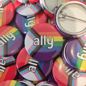 May include: A close-up of a pile of rainbow ally buttons. The buttons are round and have a silver metal backing. The buttons are all different colours of the rainbow and have the word "ally" written on them in white.
