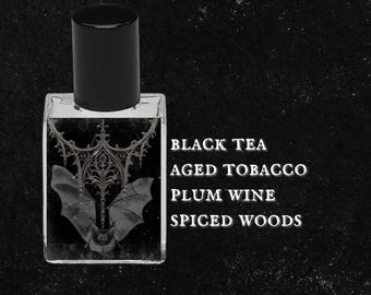 Puede incluir: Una botella de vidrio transparente con una tapa negra y una etiqueta que dice "BLACK TEA AGED TOBACCO PLUM WINE SPICED WOODS". La botella está llena de un líquido oscuro y tiene un diseño de murciélago de estilo gótico en la etiqueta.