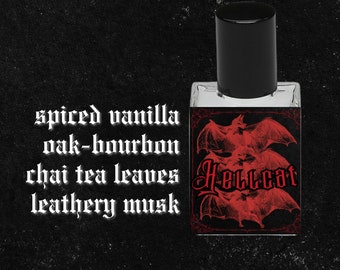 May include: A small glass bottle of perfume with a black cap. The label is black with red bats and the text "Hellcat". The scent description is "spiced vanilla, oak-bourbon, chai tea leaves, leathery musk".