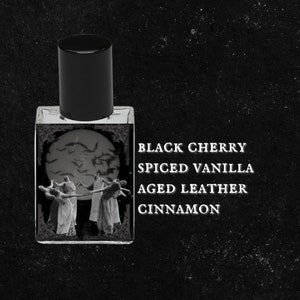 May include: A black glass bottle with a black cap and a label that reads "BLACK CHERRY SPICED VANILLA AGED LEATHER CINNAMON". The label features a black and white illustration of a group of people dancing in front of a full moon with bats flying around it.