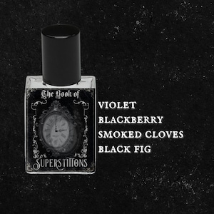 May include: A black glass bottle with a black cap and a label that reads "The Book of Superstitions". The label features a clock face and ornate details. The bottle is labeled with the scent notes: Violet, Blackberry, Smoked Cloves, Black Fig.