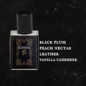 May include: A small glass bottle of perfume with a black cap and a gold label that says "Coven". The label also features a rose. The text "Black Plum, Peach Nectar, Leather, Vanilla Cashmere" is printed in white on a black background.