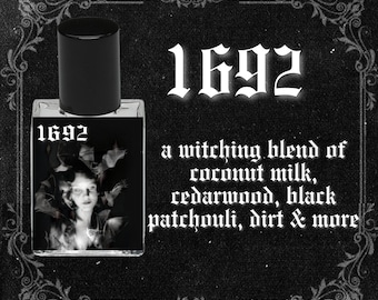 May include: A black glass bottle with a black cap and a label that says "1692". The label also has a black and white image of a woman's face with bats flying around her. The text below the image says "a witching blend of coconut milk, cedarwood, black patchouli, dirt & more".