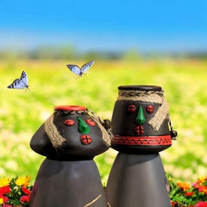 Op de afbeelding: Twee zwarte keramische sculpturen met gestileerde gezichten, versierd met rode, groene en bruine accenten. De sculpturen hebben touwdetails en staan tegen een achtergrond van vlinders en een bloemenveld.
