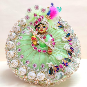 May include: A detailed, ornate doll dressed in a light green outfit with intricate embellishments. The doll features a gold face, a jeweled crown, and a matching necklace. The outfit includes white and gold accents, pink gems, and colorful feather details.