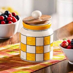 May include: A ceramic jar with a wooden lid and a white knob. The jar has a yellow and white checkered pattern. The lid is made of wood and has a white knob on top. The jar is sitting on a wooden table.