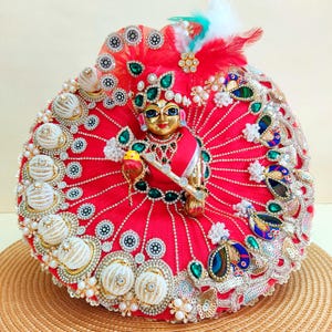 May include: A golden deity figure wearing a vibrant red dress adorned with intricate embellishments, including pearls, sequins, and emerald-colored accents. The figure holds a small bird and is topped with a feathered headdress. The overall design is ornate and detailed.