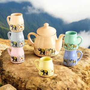 May include: A ceramic tea set featuring a beige teapot and several small mugs in pastel colors. The mugs are stacked and arranged around the teapot. Each piece is decorated with a repeating pattern of elephants and other figures.