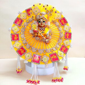 May include: A golden statue of a deity, adorned with a yellow and gold embellished outfit. The outfit features decorative elements, including floral patterns, rhinestones, and beaded accents. Small red squares with text are also visible.