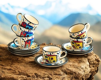 May include: A collection of small, hand-painted ceramic teacups and saucers. The cups and saucers are white with colorful house designs in red, blue, and yellow. The cups are stacked in groups of two and three.