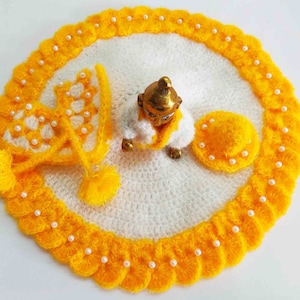 May include: A white and yellow knitted outfit for a small religious figurine. The outfit includes a dress, a hat, and a small circular mat. The outfit is decorated with white beads.