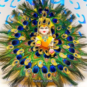 May include: A deity figurine with a yellow and gold crown and jewellery, set against a backdrop of peacock feathers. The feathers have blue, green, and gold accents. The text "Kanhaji Darbar" is visible. A decorative item.