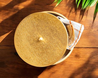 Puede incluir: Un plato redondo de color dorado con un patrón ornamental y arremolinado en la tapa. El plato está sobre una superficie de madera, con un paño a rayas blancas y naranjas y un pan plano doblado visible en el interior. Una hoja verde está en el fondo.