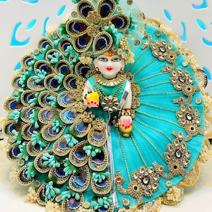 May include: A detailed figurine of a deity, adorned in a turquoise dress and elaborate peacock feather detailing. The figure has a serene expression, with gold jewelry and a small yellow object in one hand and a pink lantern in the other.