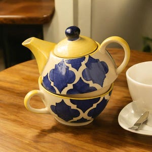 Op de afbeelding: Een keramische theeset met een theepot en een kopje. De theepot is geel en crèmekleurig met blauwe patronen. Het kopje is crèmekleurig met blauwe patronen. De set staat op een houten tafel.