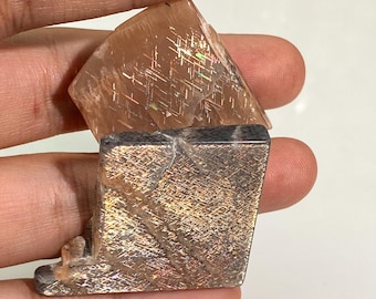 May include: Two pieces of sunstone mineral with a distinct grid-like schiller effect. The top piece is a translucent peach-colored stone, while the bottom piece is a darker, greyish-brown stone with visible internal fractures and metallic inclusions.