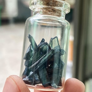 Op de afbeelding: Een klein glazen flesje met een kurkstop gevuld met donkergroene, iriserende kristallen scherven.