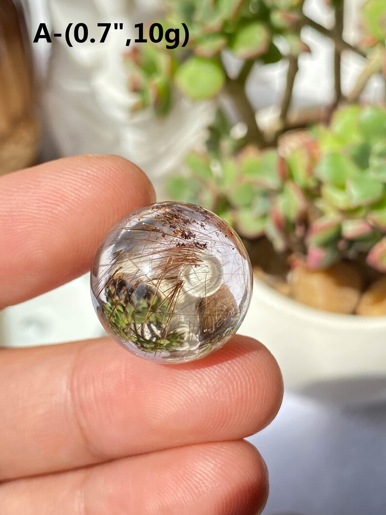 K&ouml;nnte beinhalten: Eine klare Quarzkugel mit Einschl&uuml;ssen von braunen und gr&uuml;nen haarartigen Kristallen. Die Kugel hat einen Durchmesser von etwa 1,8 cm und wiegt 10 Gramm. A- (0.7", 10g)