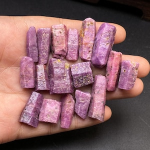 May include: A collection of raw, uncut ruby crystals in varying shades of pink and purple. The gemstones are held in a hand, showcasing their natural, rough textures and crystalline structures. The crystals range in size and shape.
