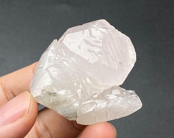 Puede incluir: Un grupo de cuarzo cristalino transparente con múltiples facetas. El cuarzo es de un color pálido y translúcido, con una textura ligeramente rugosa. El grupo se sostiene en una mano sobre un fondo oscuro.