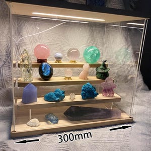 Könnte beinhalten: Eine Holz- und Acryl-Vitrine mit LED-Beleuchtung, die eine Sammlung von Kristallen und Kugeln zeigt. Die Vitrine enthält Objekte in Rosa, Blau, Grün und Klar. Abmessungen: 27,3 cm x 30 cm.