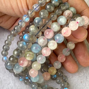 May include: Several beaded bracelets featuring a variety of gemstone beads. The beads are primarily round and come in shades of gray, pink, blue, and clear. The bracelets are strung on a clear elastic cord and are held in a hand.