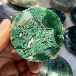 Peut inclure: Une pierre d'agate mousse polie, de forme circulaire, pr&eacute;sentant un motif vert, blanc et clair saisissant. La pierre est tenue dans une main, avec d'autres pierres similaires en arri&egrave;re-plan. L'agate a un aspect naturel et organique.