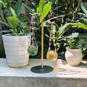 May include: A gold-coloured earring display stand with two pairs of earrings. One pair has a green sphere, the other a yellow sphere, both in gold wire and purple beads. The stand is on a dark green base, with plants in the background.