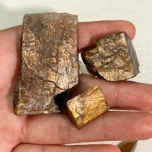 Op de afbeelding: Drie ruwe zonnesteen mineralen met een metallic, glinsterende goudbruine glans. De stenen hebben een blokvormige, kristallijne structuur met zichtbare strepen en reflecterende insluitingen die het licht vangen, wat een warme, aardse kleurenpalet laat zien.