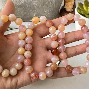 Puede incluir: Una hebra de cuentas de piedras preciosas de color rosa, naranja y blanco. Las cuentas son redondas y tienen un acabado liso y pulido. Las cuentas están enhebradas en un cordón elástico transparente.