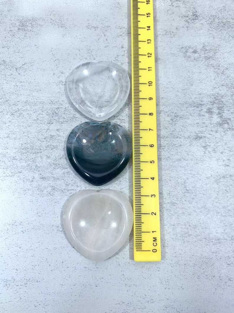 May include: Three heart-shaped crystal bowls, one clear, one black and white agate, and one pink, with a yellow ruler for size comparison.