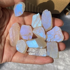 May include: A collection of raw moonstone crystals, each with a unique shape and a pearly, iridescent sheen. The stones display flashes of blue and white, held in a hand against a soft, neutral background.