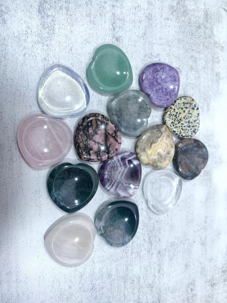 May include: A collection of 12 heart-shaped crystal bowls in various colors, including green, purple, pink, and clear. The bowls are arranged in a circle on a light gray surface.