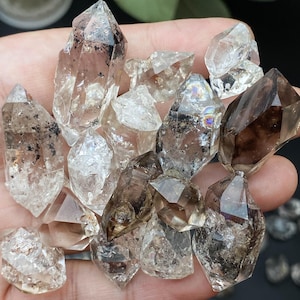 Puede incluir: Un puñado de cristales de cuarzo transparente y ahumado. Los cristales varían en tamaño y forma, con puntas afiladas e inclusiones naturales. Los colores van del transparente al marrón claro, algunos con áreas oscuras y ahumadas. Los cristales se sostienen en una mano.
