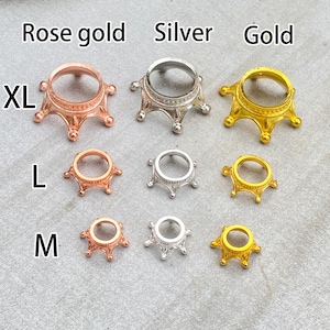 Può includere: Una collezione di supporti decorativi in metallo color oro rosa, argento e oro. I supporti sono disponibili nelle misure XL, L e M. I supporti hanno un design a forma di corona e sono esposti su una superficie grigia.