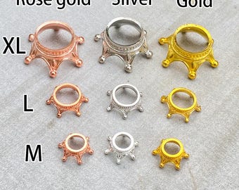 May include: A selection of decorative metal stands in rose gold, silver, and gold. The stands are available in XL, L, and M sizes. The stands have a crown-like design and are displayed on a grey surface.