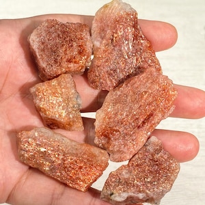 Può includere: Una collezione di sei esemplari di pietra del sole grezza caratterizzati da una tonalità rosso-arancio con inclusioni metalliche scintillanti. Queste pietre grezze e non lucidate presentano una consistenza cristallina e forme angolari.